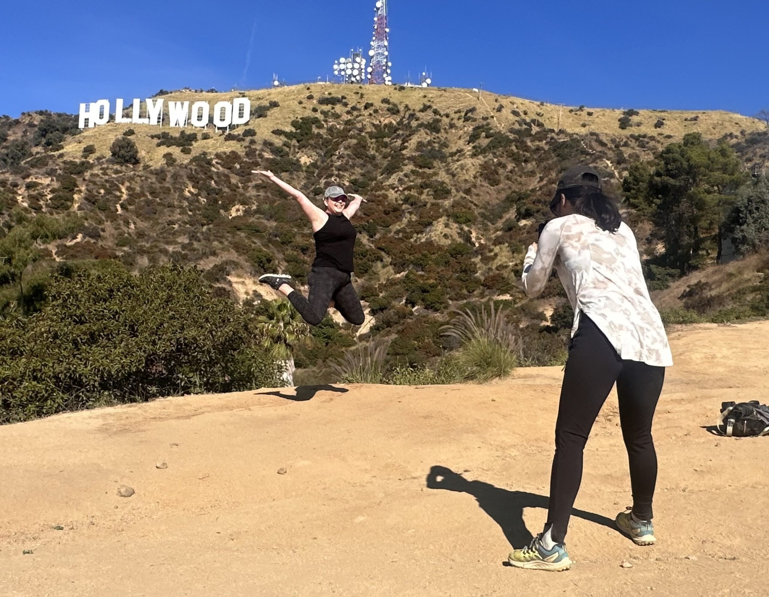 A girl takes a photo of another girl jumping in front of the Hollywood Sign on the Hollywood Sign Tour